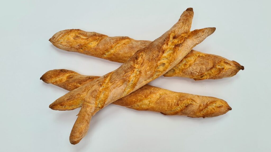 Zwei klassische Baguettes "Pain de Tradition", auf denen quer ein Baguette Sarmentine liegt; das Baguette Sarmentine hat jeweils zwei spitze Enden anstatt eines