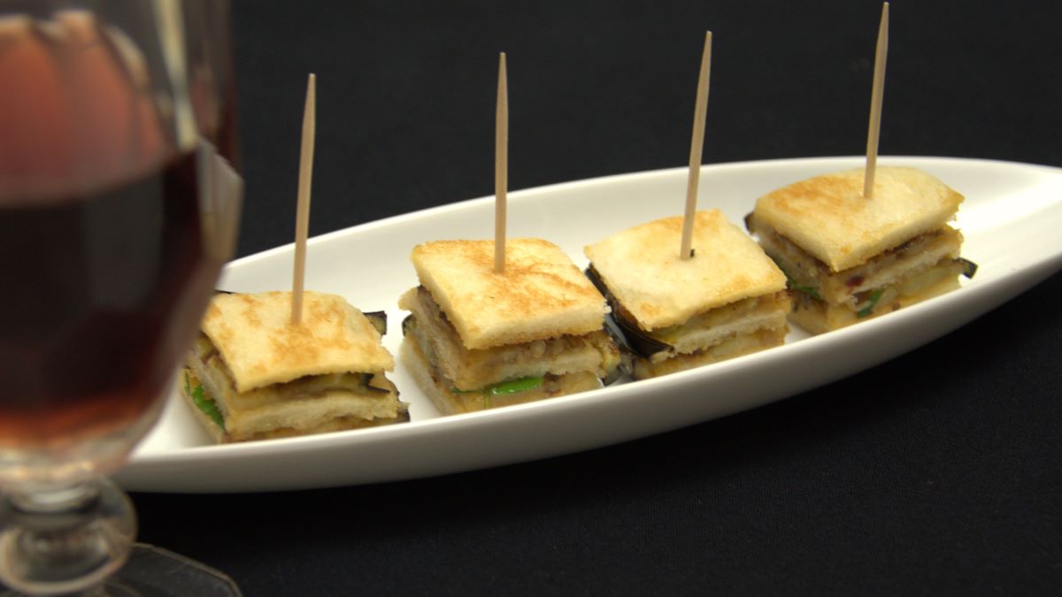 Auberginen-Millefeuille: vier geschichtete Häppchen mit geröstetem Tramezzinibrot-Scheiben, dazwischen gebratene Auberginen und verschiedene Gewürze und Chutneys. Angerichtet auf einem weißen Porzellanschälchen in Bootsform.