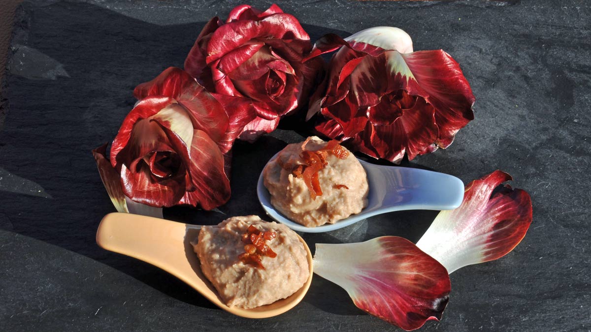 Rosa von Gorizia mit Borlottibohnen: Dieses Bild zeigt eine Variante dieser Kombination. Auf einem schwarzen Granitbrett liegen ganze Rosen von Gorizia, ein paar lose Blätter sowie zwei Servierlöffel mit einem kalten Bohnenpüree, auf dem knusprige Speckstückchen angerichtet sind.