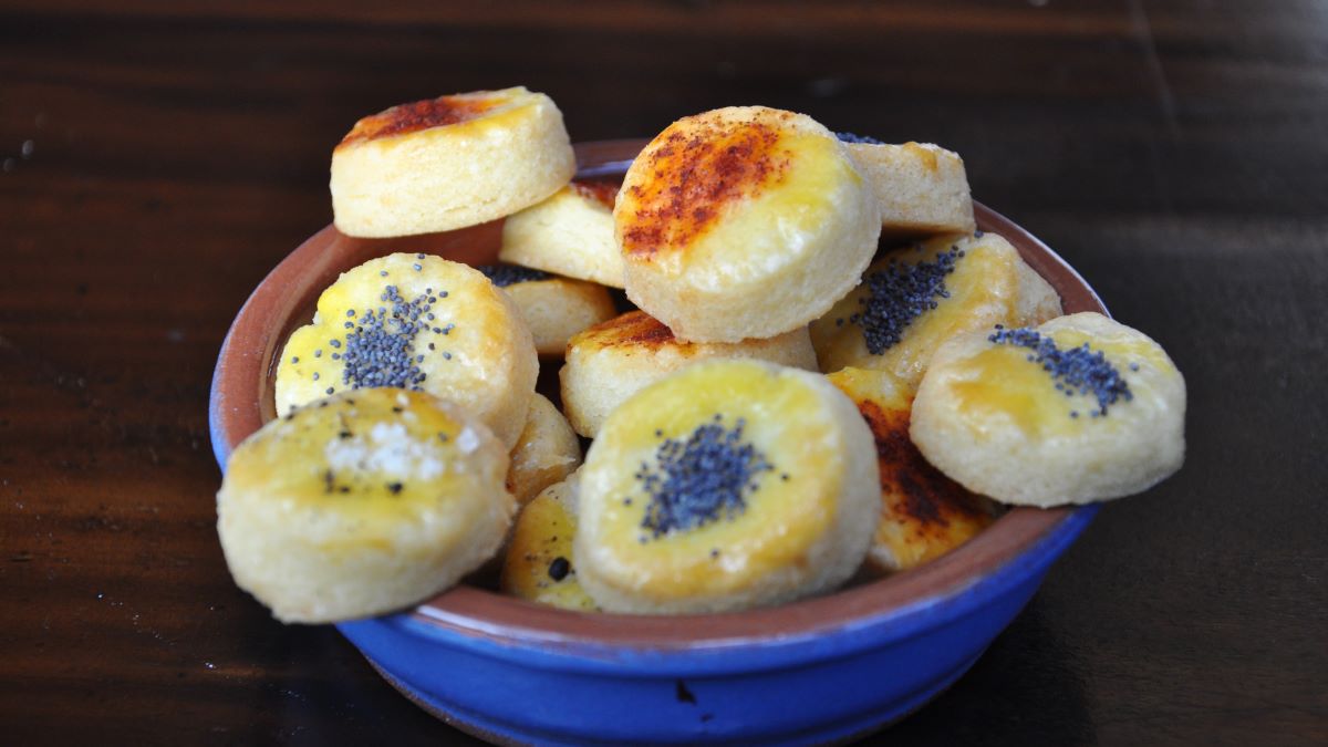 Das Bild zeigt eine kleine Schüssel in Großaufnahme, in der verschieden bestreute Sablés au Gruyère präsentiert werden: rot mit Paprika, schwarzblau mit Mohn und weiß mit grobem Salz