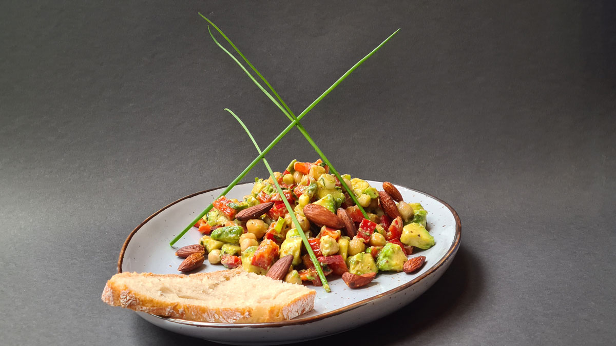 Auf dem Bild: Ein wärmender Wintersalat auf einem Teller vor grauem Hintergrund. Die Zutaten Avocado, Mandeln, Paprika, Kichererbsen sind gut zu erkennen, darüber sind einige Halme Schnittlauch gesteckt, die den Salat in die Höhe führen