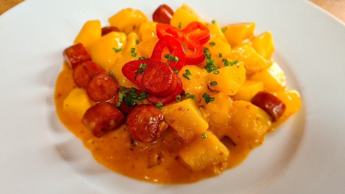 Ein farbenfrohes Wiener Erdäpfelgulasch: Zu sehen sind Kartoffelwürfel, Wurstscheiben, rote Paprikascheiben, Schnittlauch und Saft auf einem weißen Teller.