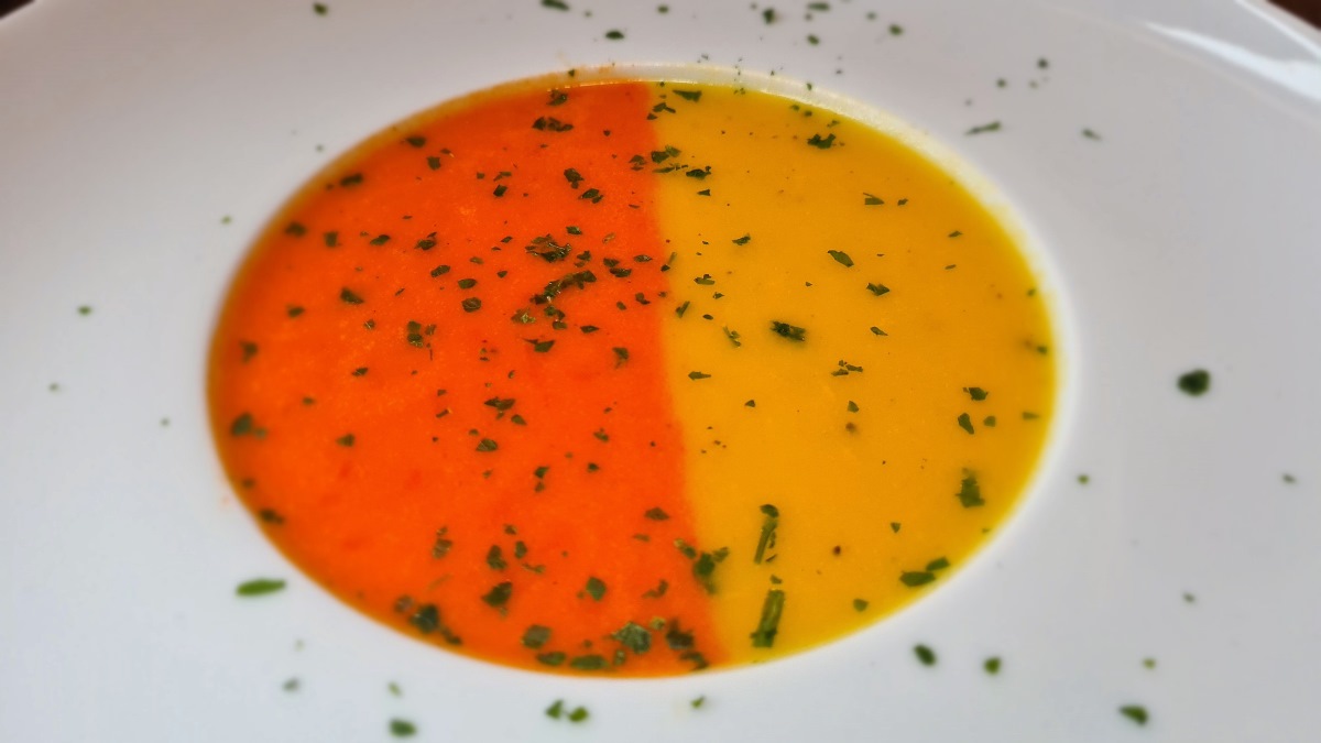 So sieht die zweifärbige Paprikasuppe aus. In einem weißen Suppenteller befindet sich die angerichtete Suppe, links die orange, rechts die gelbe Version. Darauf gehackte Petersilie.