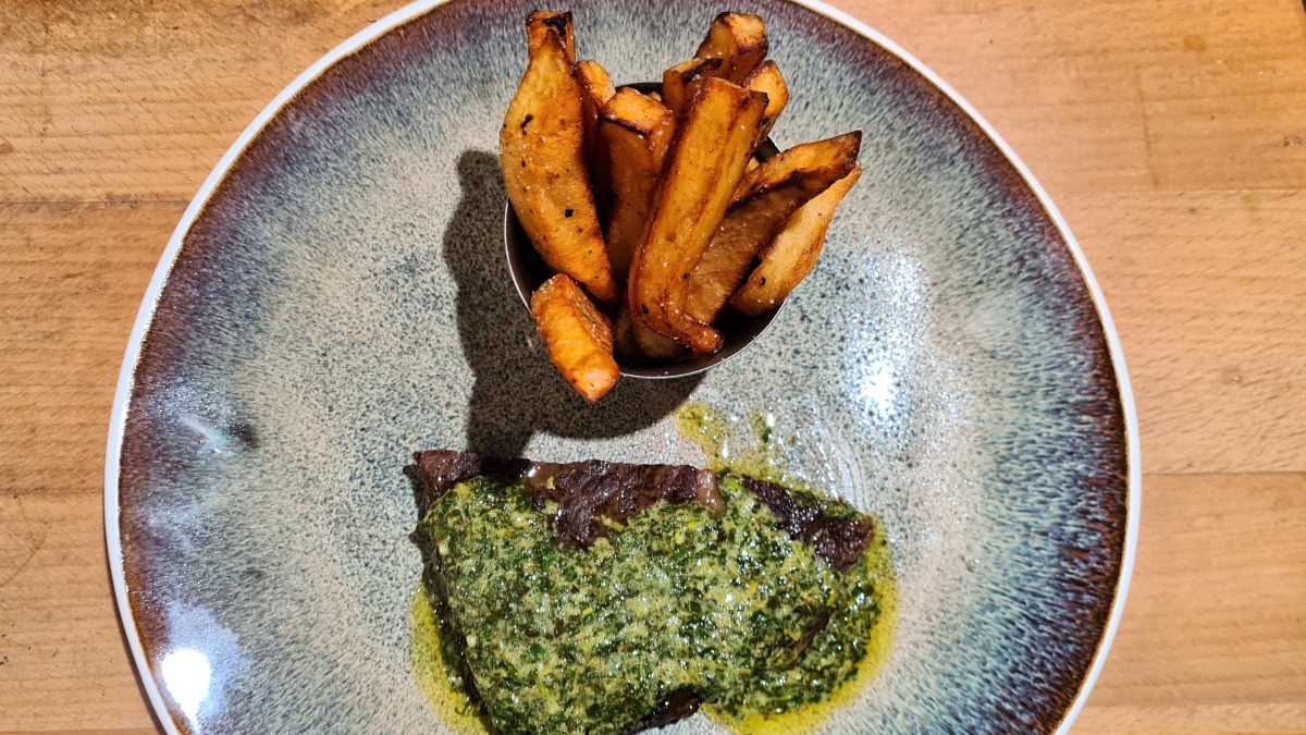 Auf dem Teller ein Entrecôte Double „Café de Paris“, daneben eine Schale mit Pommes frites. Die Buttersauce „Café de Paris“ liegt auf dem Steak, sie ist von den Kräutern grün.