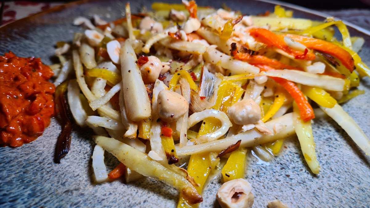 Warmer Wurzelsalat in Großaufnahme: Deutlich zu sehen die Vielfalt der Gemüsesorten, der Wurzeln und Knollen wie Pastinake, Gelbrübe, Karotte, darüber Haselnüsse und daneben roter Ajvar.