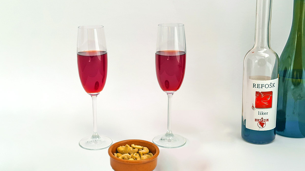 Kir Karst - der neue Aperitif: Es sind zwei Sektgläser zu sehen, der Cocktail darin ist rot. Rechts daneben eine Flasche Refosko-Likör des Weinguts Hrvatin.