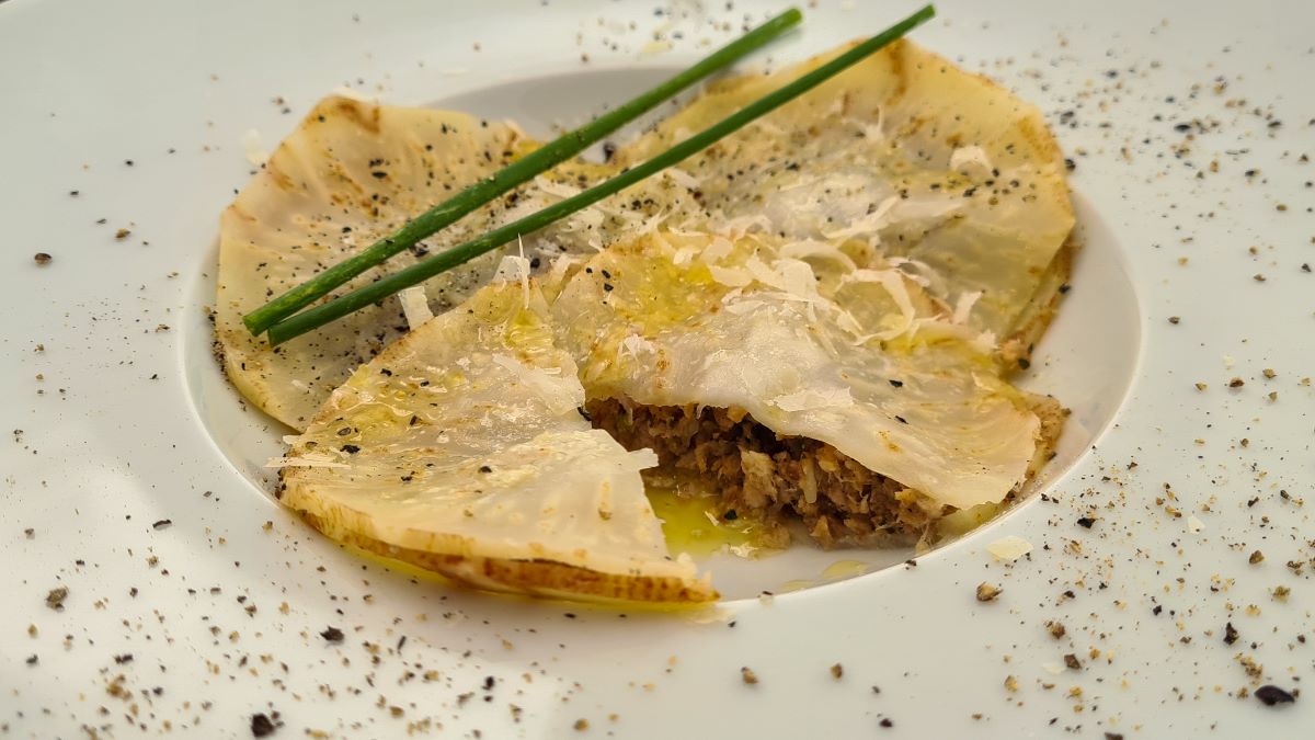 Auf dem Bild sind drei Zeller-Ravioloni zu sehen, darüber geriebener Parmesan und Schnittlauchhalme. Ein Raviolono ist aufgeschnitten, damit die Fülle darin sichtbar ist.