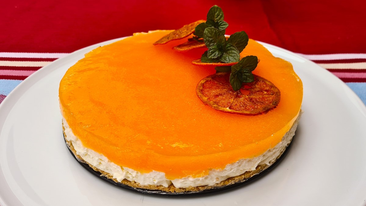 Eine Aperol-Spritz-Torte auf einem weißen Tortenteller. Die Aufnahme zeigt den knusprigen Boden, die Prosecco-Creme und die Aperol-Orangen-Auflage. Darauf gedörrte Orangenscheiben und Minzezweige als Deko