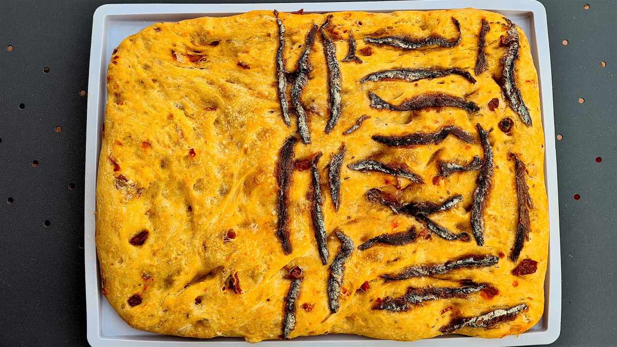 Aromatische Focaccia auf dem Blech. Eine Hälfte ist mit Anchovis belegt, auf der anderen Hälfte sieht man die Tomatenstücke im Teig besser.