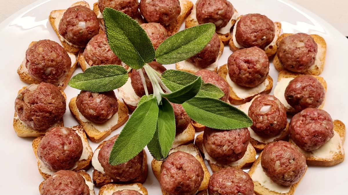 Ein Teller mit Beef Tatar als kleiner Genuss: Man sieht Minizwiebackscheiben bestrichen mit Butter und belegt mit einer kleinen Kugel Beef Tatar. Darüber ein ganzer Salbeizweig