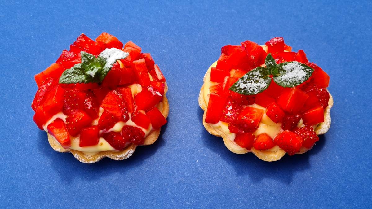 Zwei Erdbeertörtchen auf blauem Untergrund; die gelbe Crème pâtissière, darauf die Erdbeerwürfelchen, bestreut mit Staubzucker und einem Minzblättchen
