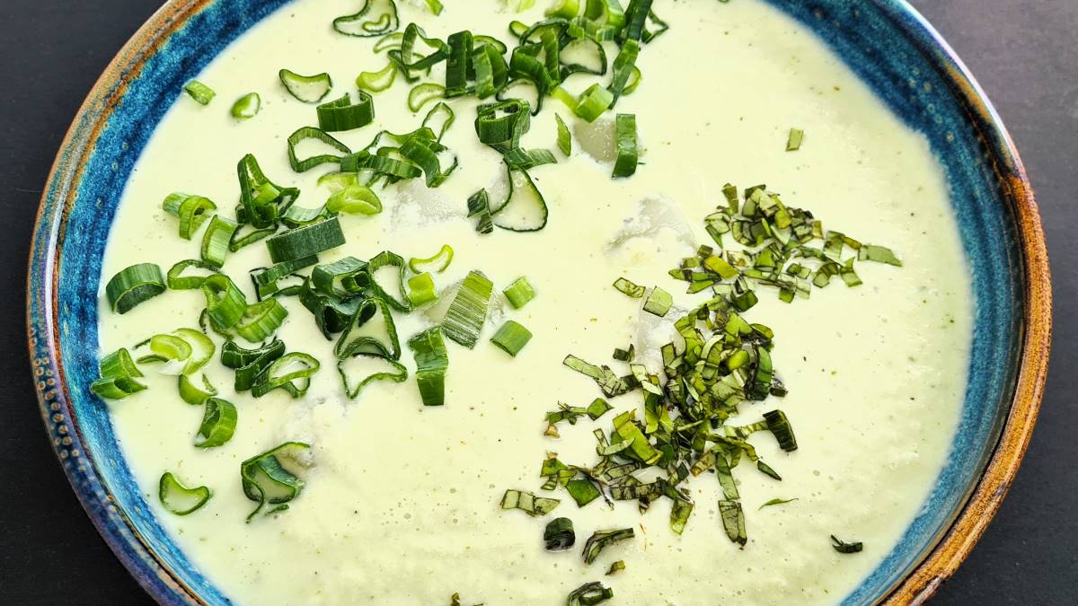 In einem Supenteller ist die kalte Basilikumgurkensuppe angerichtet, darauf sind grüne Röllchen vom Jungzwiebel auf der einen Hälfte der Oberfläche und auf der anderen gehacktes Basilikum