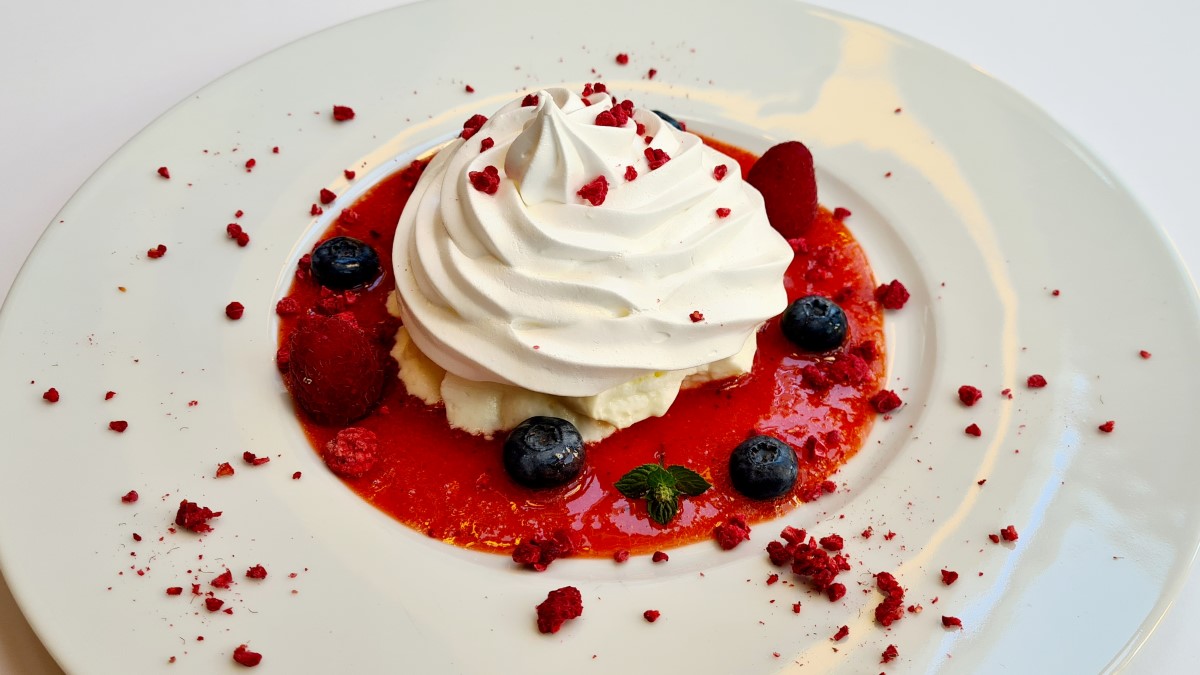 Gefülltes Baiser, hier auf einem roten Fruchtspiegel, frischen Beeren und gefriergetrockneten Himbeeren auf einem weißen Teller