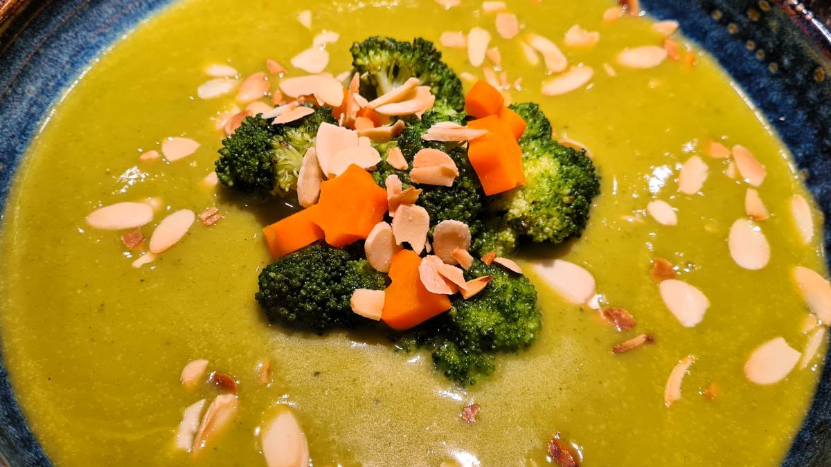 Das Bild zeigt die einfache Brokkolicremesuppe in einem blauen Suppenteller. Auf der Suppe sind Brokkoli-Röschen, Mandelsplitter und Karottensternchen zur Deko angerichtet.