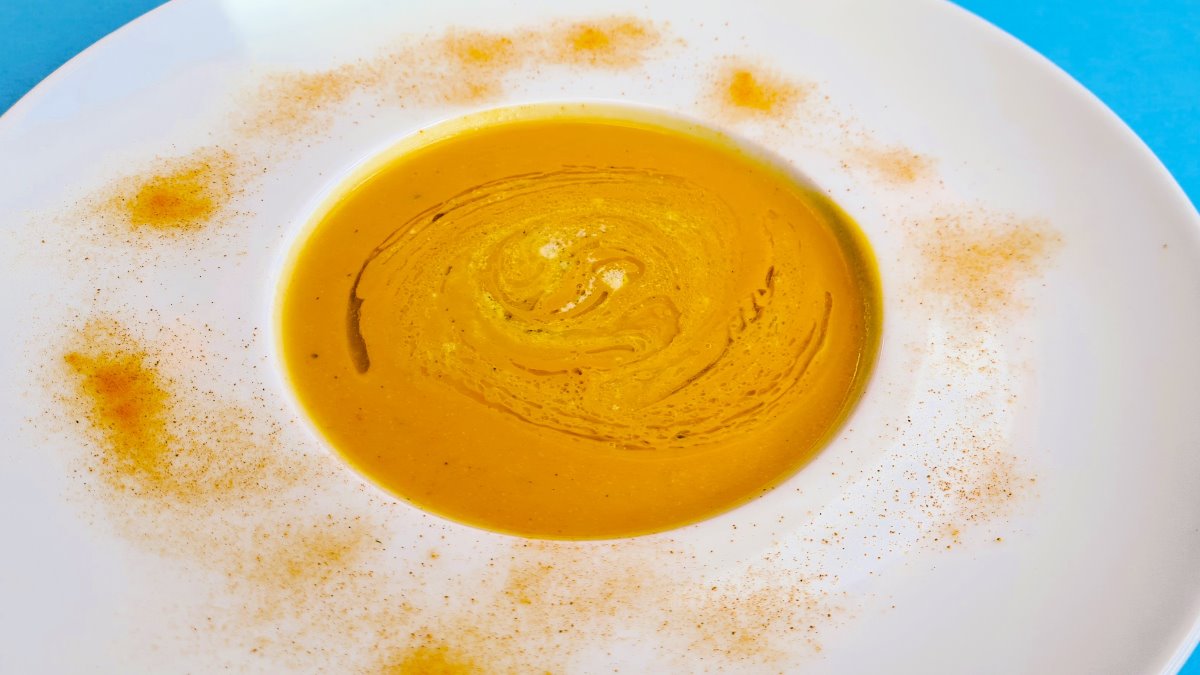 Fruchtige Kürbissuppe: Man sieht einen Suppenteller mit einem breiten Rand, die goldgelbe Suppe in der Mitte, darauf Orangenöl und geschmolzener Blauschimmelkäse, auf dem Tellerrand Kurkumapulver zur Deko