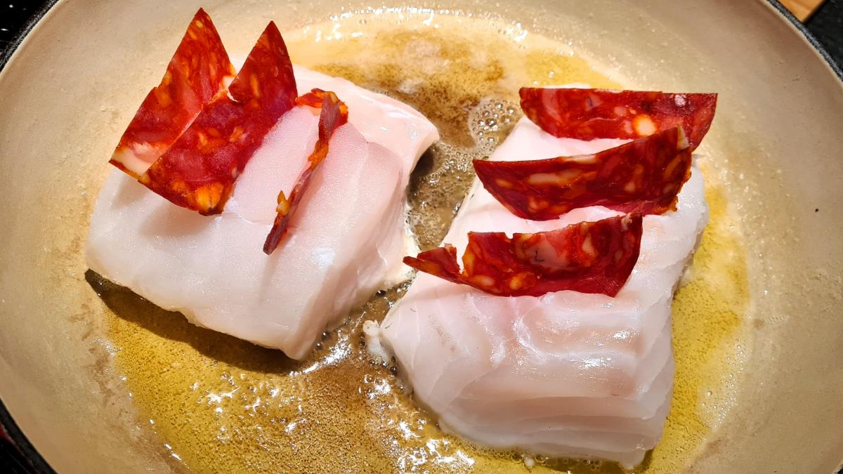 Die Kabeljaurücken mit Chorizo gespickt in der Pfanne, man sieht das heiße Öl und das noch rohe Fischfilet