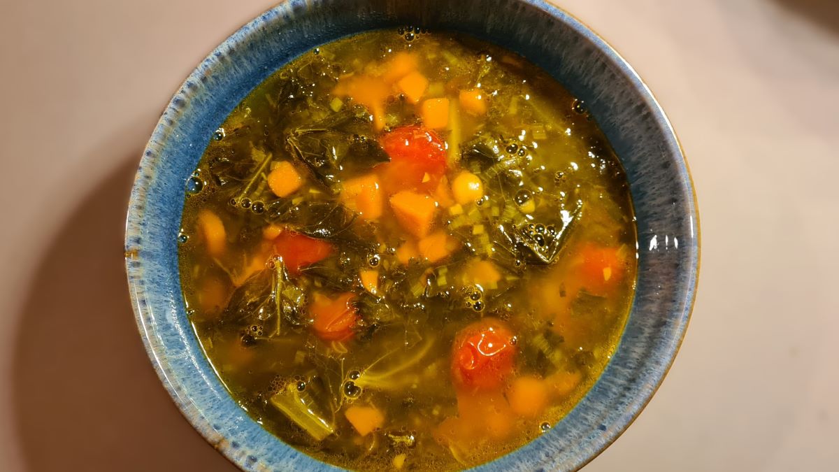 Kürbis-Grünkohl-Minestra: Das Bild zeigt die Suppe in einer blauen Suppenschale, in der die Zutaten gut zu erkennen sind: Kürbisstücke, Grünkohlblätter, Kichererbsen, Tomaten und Gewürze.