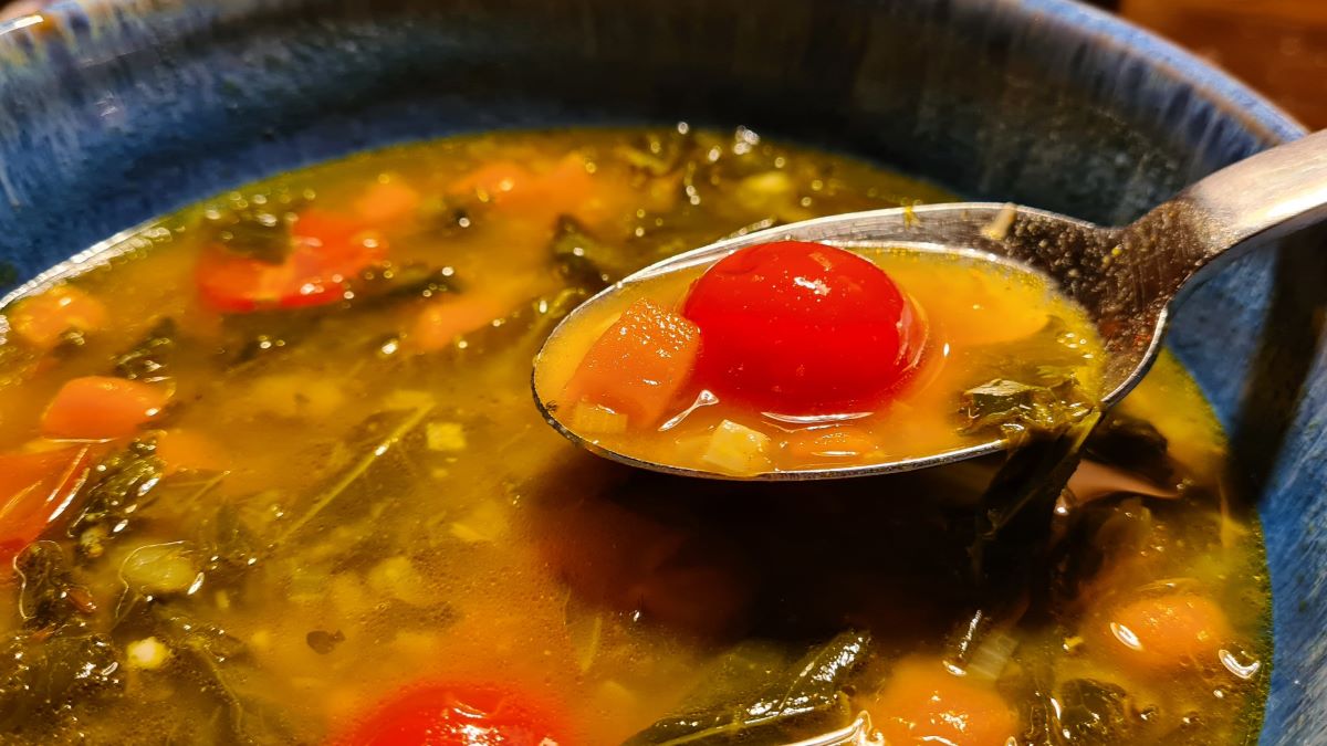 Kürbis-Grünkohl-Minestra: Das Bild zeigt die Suppe in einer blauen Suppenschale mit einem Löffel, auf dem eine Tomate, Kürbis- und Grühnkohlstücke zu sehen sind. In der Suppe sind die Zutaten gut zu erkennen.