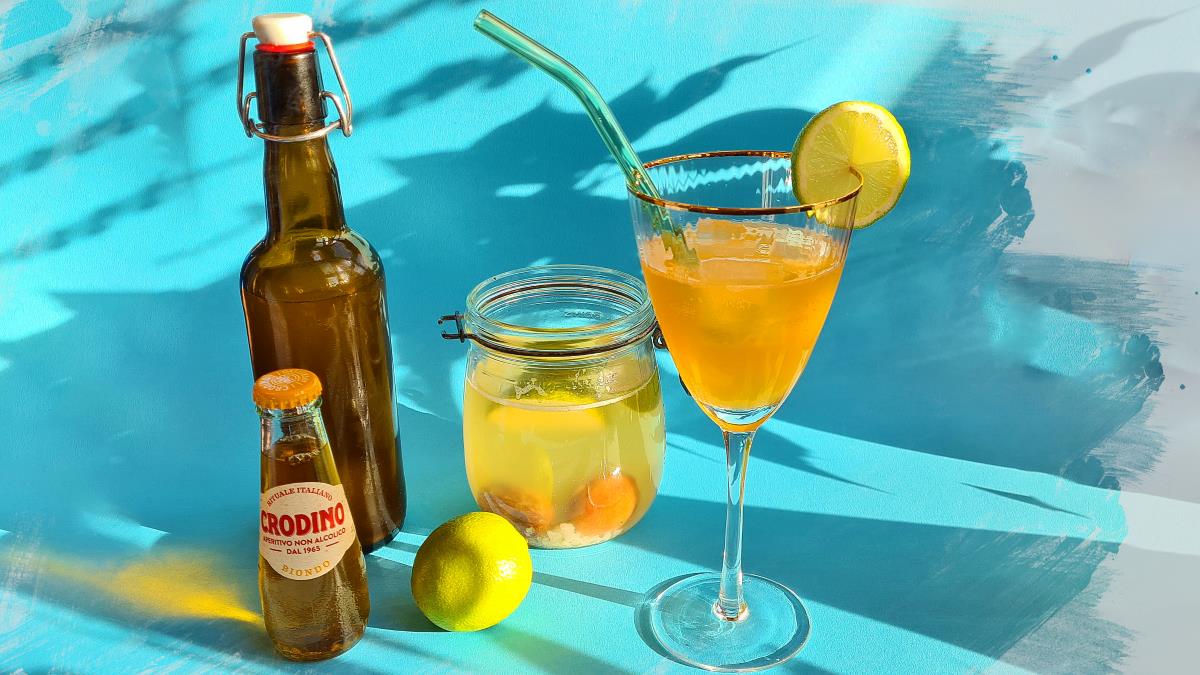 Das Bild zeigt den Drink und die Zutaten von einem Limetten-Kefir-Spritz: Auf blauem Hintergrund sind eine Flasche Wasserkefir, der Wasserkefiransatz im Glas, eine Limette, ein Fläschchen gelber Crodino und der Drink selbst zu sehen. Dieser ist mit einer Limettenscheibe und einem grünen Glashalm dekoriert.