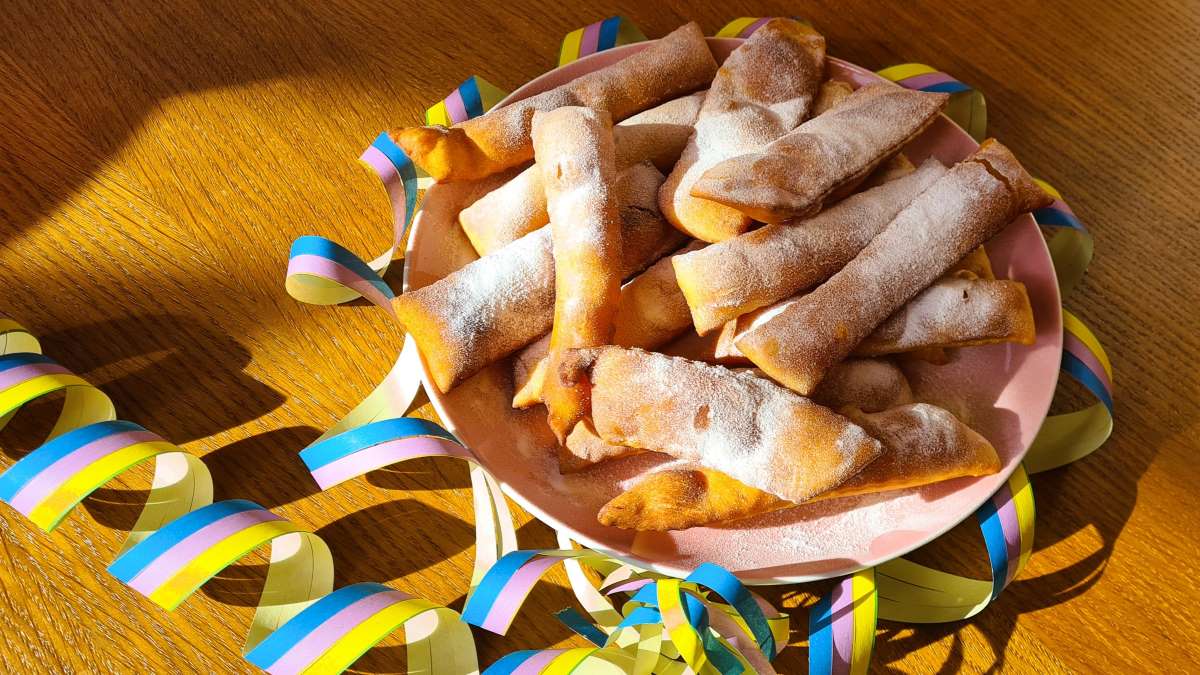 Das Bild zeigt einen Teller Karnevalsgebäck Crostoli, mit Staubzucker überstreut und umgeben von Faschingsgirlanden.