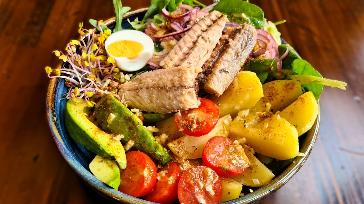 Das Bild zeigt eine wunderschön bunt angerichtete Bowl La Rochelle auf einem braunen Holztisch in einer blauen Keramikbowl. Die Zutaten sind übersichtlich angeordnet: Hartes Ei, Thunfisch, Avocado, gekochte Kartoffeln, Kirschtomaten, Sprossen, Zwiebel und Blattsalat.