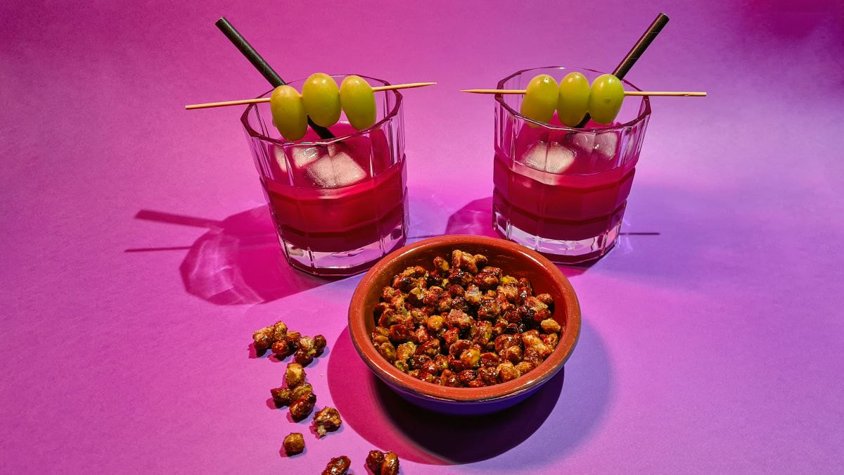 Orientalischer Aperitif: Das Bild zeigt auf einem violetten Grund den alkoholfreien roten Drink aus Verjus, Rosensirup und Granatapfelsaft, auf dem Glasrand hängen grüne Weintrauben als Deko, sowie eine Schale mit Pistazien, die mit Salzkaramell ummantelt sind.