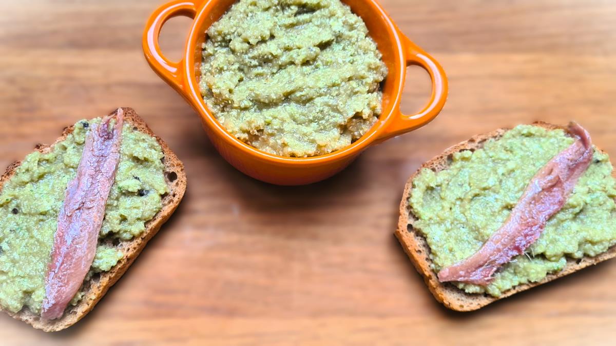Das Bild zeigt den Puntarella-Aufstrich in einem orangen Töpfchen und aufgetragen auf zwei Brötchen, die mit einer eingelegten Sardelle belegt sind.