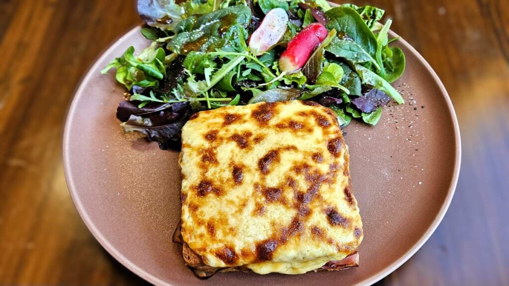 Croque Monsieur: Das Bild zeigt den angerichteten Croque Monsieur auf einem braunen Teller, daneben Blattsalat mit Dressing und rot-weißen Radieschen