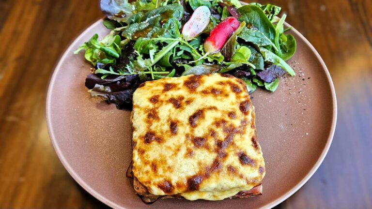 Croque Monsieur: Das Bild zeigt den angerichteten Croque Monsieur auf einem braunen Teller, daneben Blattsalat mit Dressing und rot-weißen Radieschen