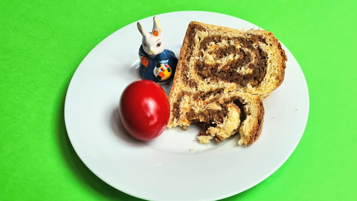Das Bild zeigt ein Stück Gubana auf einem weißen Teller mit einem roten Osterei einem Osterhasen aus Keramik vor grünem Hintergrund. Gut erkennbar ist die reiche Füllung des Gebäcks.