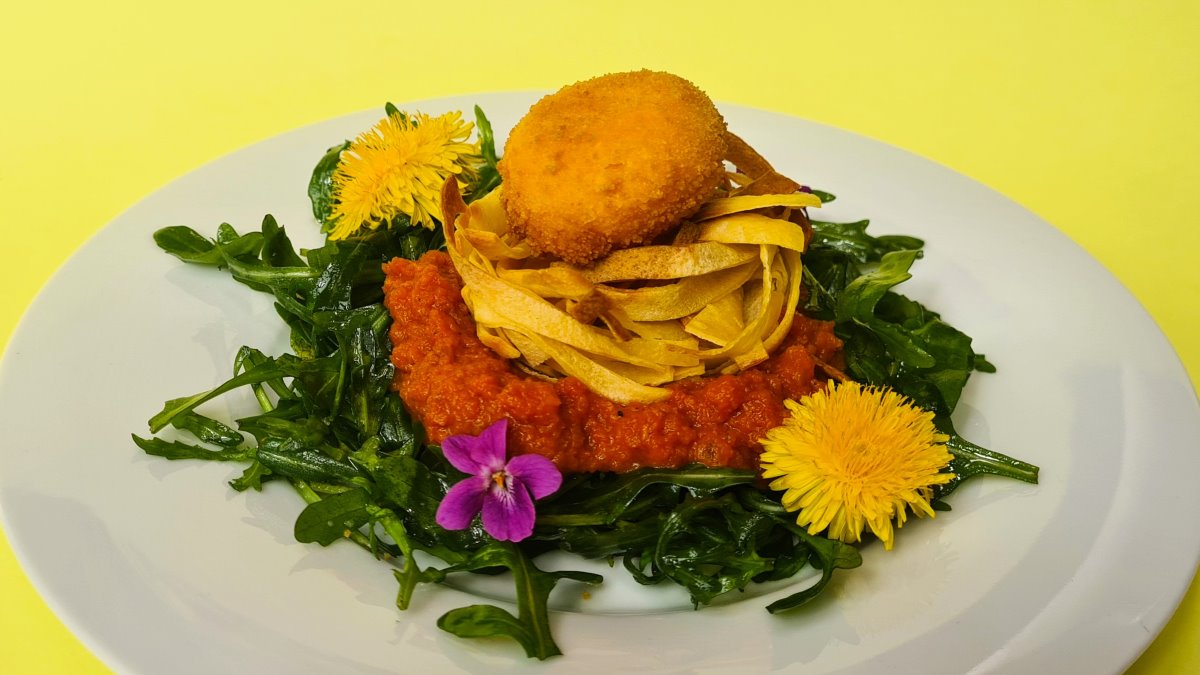 Das Bild zeigt auf einem weißen Teller marinierte Rucolablätter, darauf eine Tomatensauce, darauf knusprige Fritatten und darauf ein frittierter Dotter. Dokoriert ist das Gericht mit essbaren Blüten, hier Veilchen und Löwenzahn.