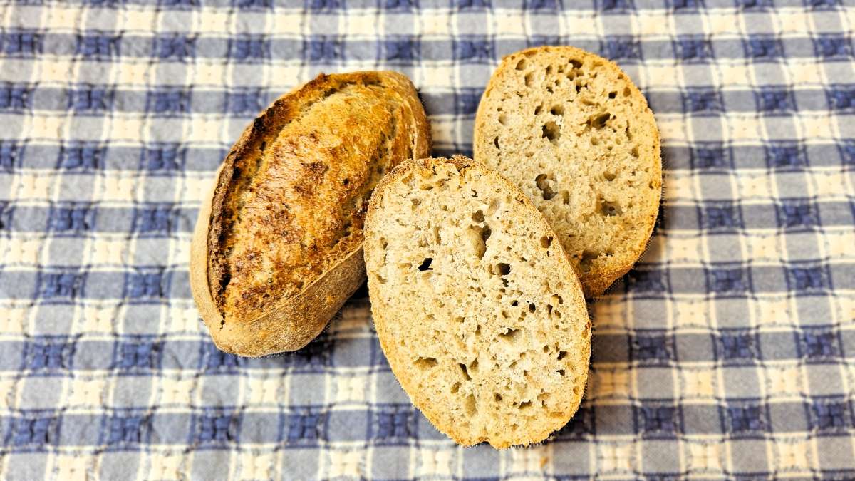 Dinkelkeimlinge: Das Bild zeigt auf einem blau karierten Tuch zwei Weckerln, eines längs aufgeschnitten, damit die Krume sichtbar ist. Links zeigt das ganze Brötchen seine rösche Kruste.