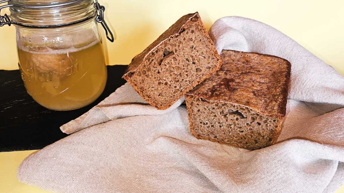 Kefir-Dinkel-Vollkornbrot auf einem Tuch im Anschnitt: das Bild zeigt die dunkle, unregelmäßige Krume der wilden Hefen aus dem Wasserkefir, dessen Ansatz in einem Fermentier-Glas daneben steht, mit Trockenfeige und Zitronenscheibe.