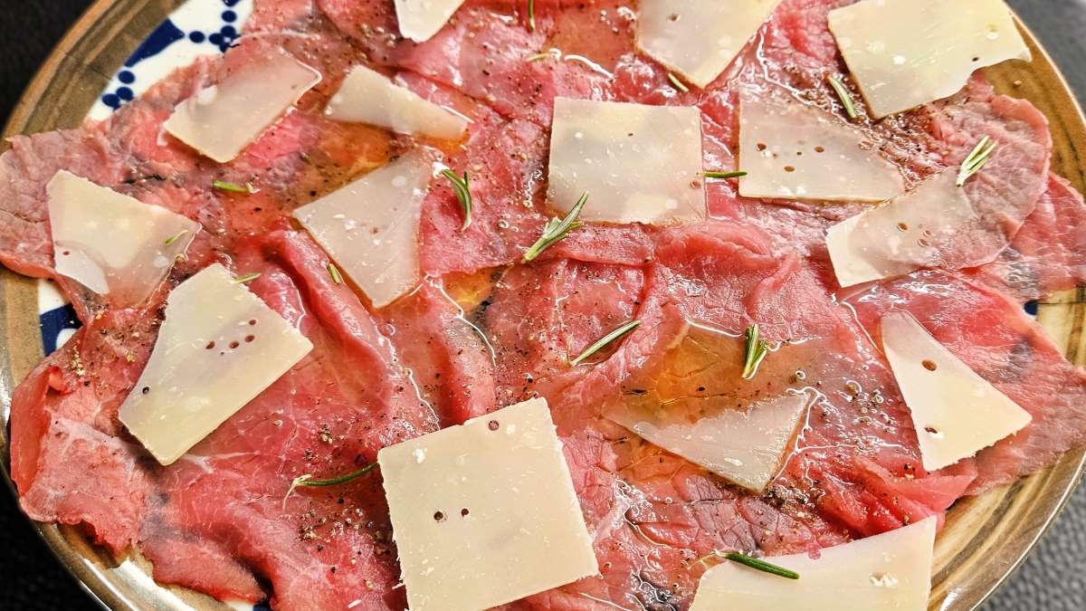 Carpaccio di Manzo puristisch: Auf dem Teller liegen die dünnen Fleischscheiben, darauf Öl, Parmesanhobel und Rosmarin