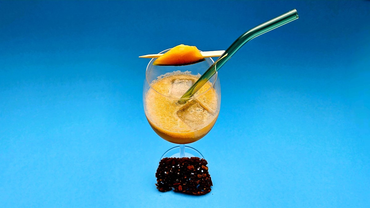Pineau Melon Pep’s: Auf einer blauen Fläche präsentiert sich der Drink in einem wunderschönen Dunkelgelb, dazu ein Melonenstückchen als Deko und ein Glasstrohhalm. Am Fuß des Glases liegt ein Brotchip.