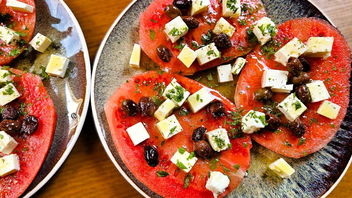 Wassermelonensteaks mit Feta, auf dem Bild zwei Teller mit dunkelblauen-grauen Design, auf denen je drei ganze Scheiben der Minimelone angerichtet sind, darauf Feta und Oliven, Kräuter und das Calamansi-Dressing
