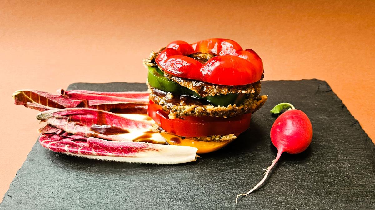Grünkern-Millefeuille, das Bild zeigt die Schichten aus grünem und rotem Paprika sowie den Scheiben aus dem Grünkern-Laibchen, links liegen rote Chicorée-Blätter, rechts ein Radieschen.