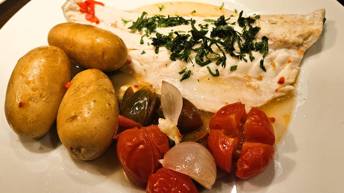 Fischfilet in Acqua Pazza: Das Bild zeigt das angerichtete Fischfilet mit dem Gemüse und Kartoffeln auf einem weißen Teller. Das Filet ist mit gehackten Kräutern bestreut und leicht mit dem Sud übergossen.