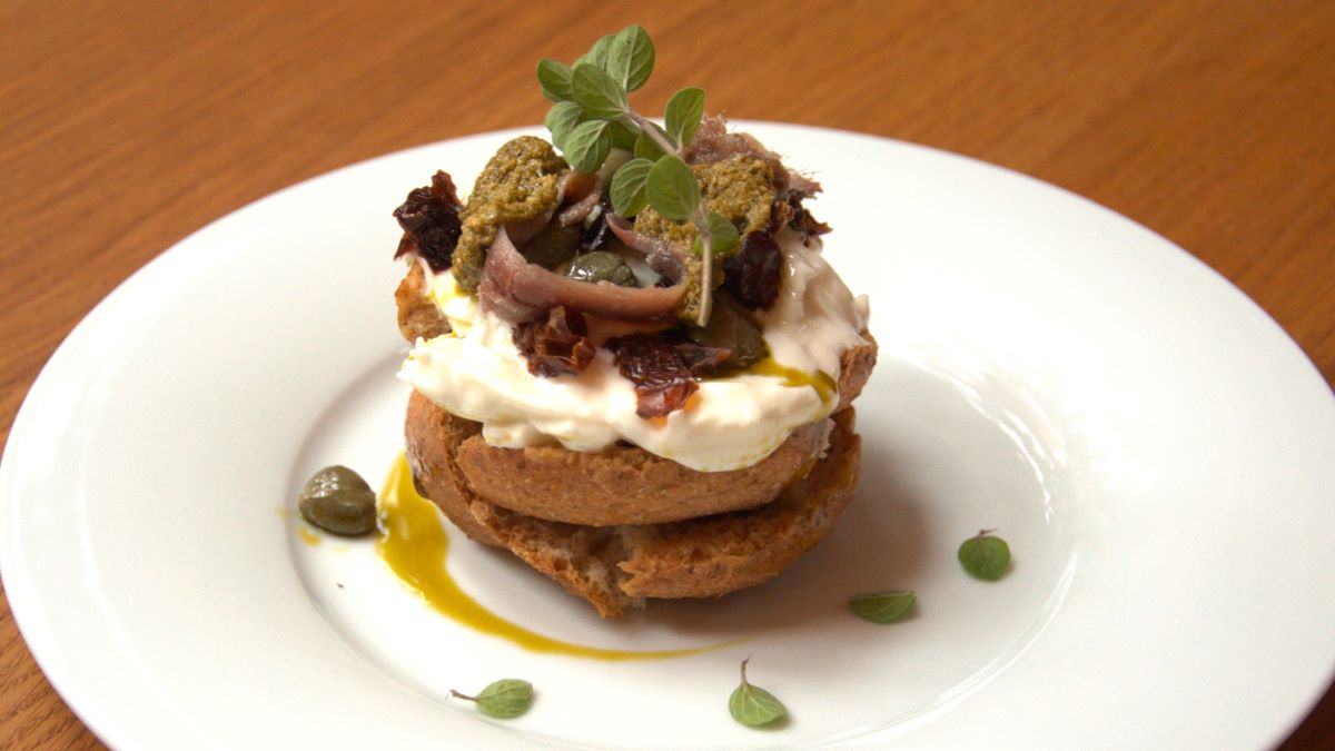 Antipasto mit Friselle: man sieht von oben die Schichten von den Friselle-Scheiben, Stracciatella, Pesto, Kapern, Sardellen, Gewürze und Kräuter auf einem weißen Teller angerichtet