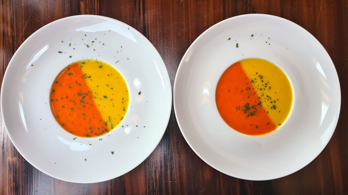 Zweifärbige Paprikasuppe: In zwei weißen Suppentellern befindet sich die angerichtete Suppe, die linke Hälfte orange, die recht gelb. Darauf gehackte Petersilie.