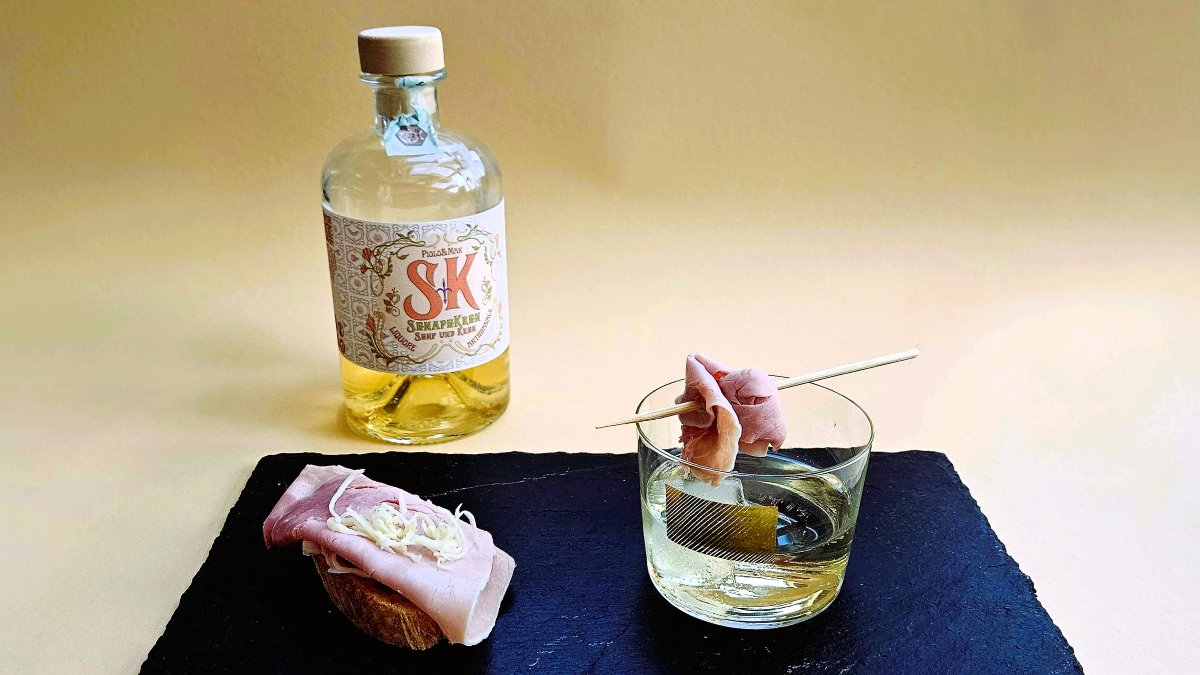 Das Bild zeigt den Senf-Kren-Likör aus Triest, im Hintergrund die Flasche, davor auf einem schwarzen Schieferbrett links ein Brötchen belegt mit Schinken, darauf geriebener Kren (Meerrettich), recht das Glas des Drinks, darauf auf einem Spieß der Schinken als Deko