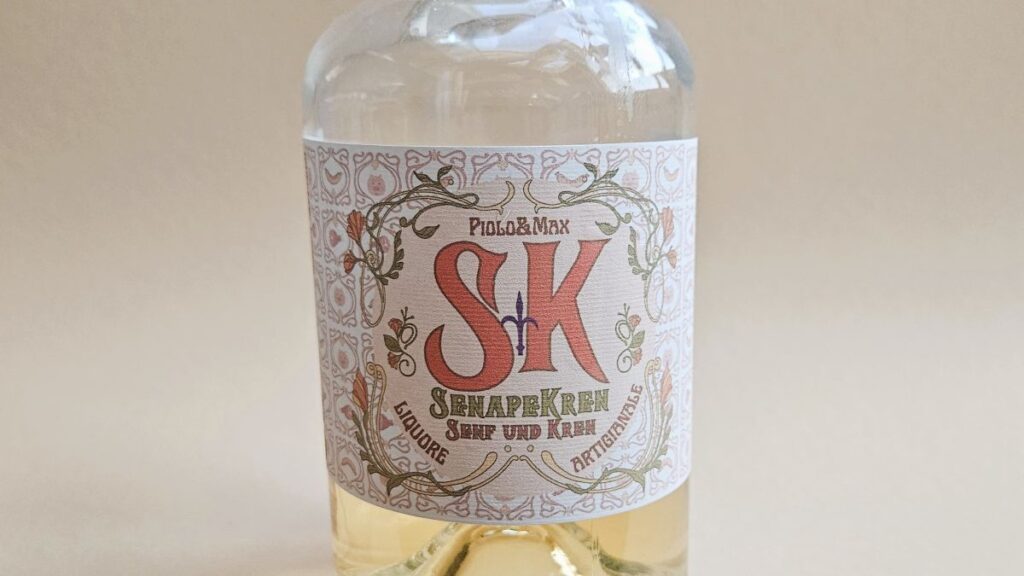 Das Bild zeigt das Etikett der Flasche vom Senf-Kren-Likör aus Triest: Das Design bezieht sich auf den Liberty-Stil, es steht darauf: Piolo&Max, S/K Senape e Kren, Senf und Kren, Liquore Artigianale