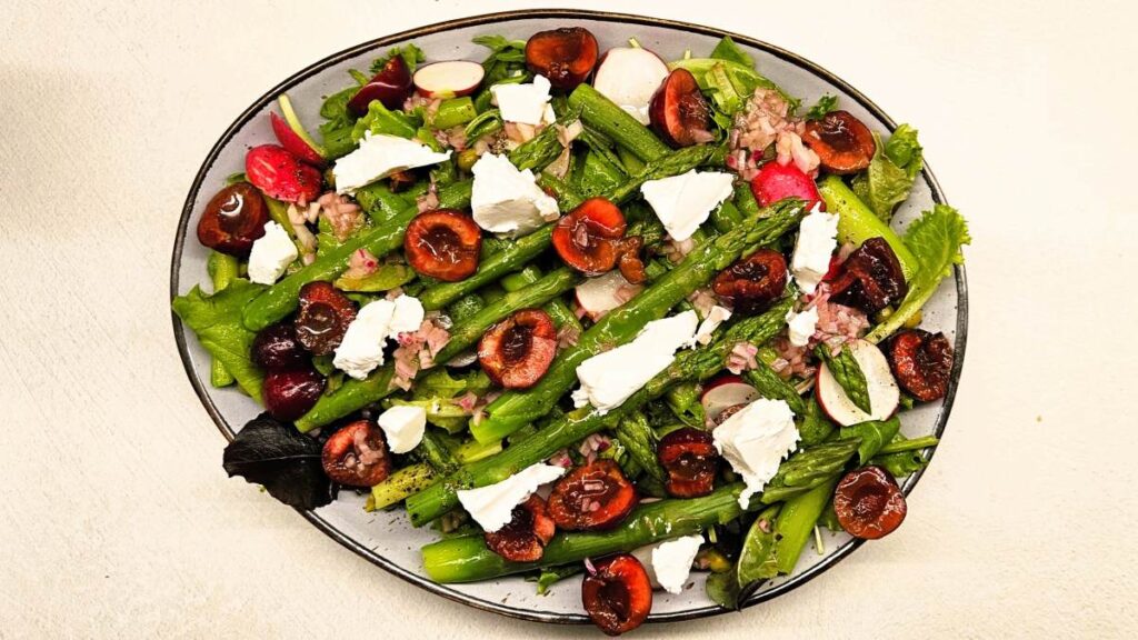 Auf dem Teller in Draufsicht ist der Kirschensalat mit Spargel angerichtet. Zu sehen sind die Zutaten: grüner Spargel, rote halbierte Kirschen, darunter gemischter Blattsalat und vereinzelt Erbsen. Über dem Salat sind weiße Frischkäsestücke.