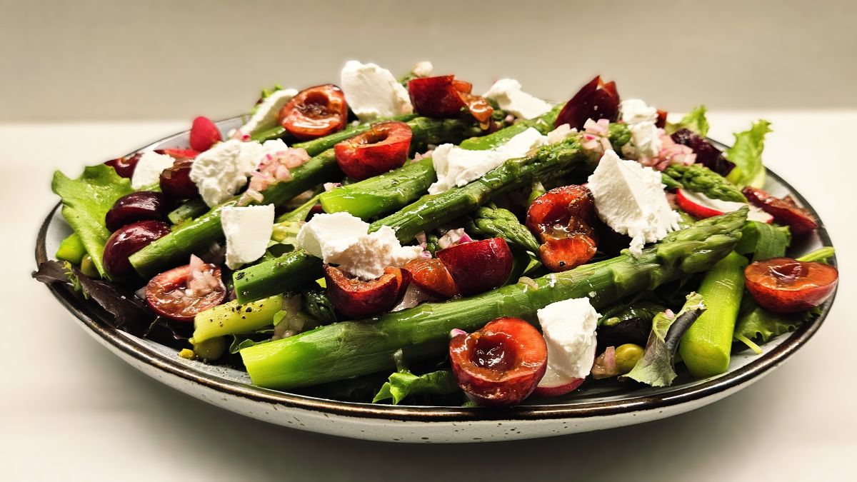 Auf dem Teller ist der Kirschensalat mit Spargel angerichtet. Zu sehen sind die Zutaten: grüner Spargel, rote halbierte Kirschen, darunter gemischter Blattsalat und vereinzelt Erbsen. Über dem Salat sind weiße Frischkäsestücke.