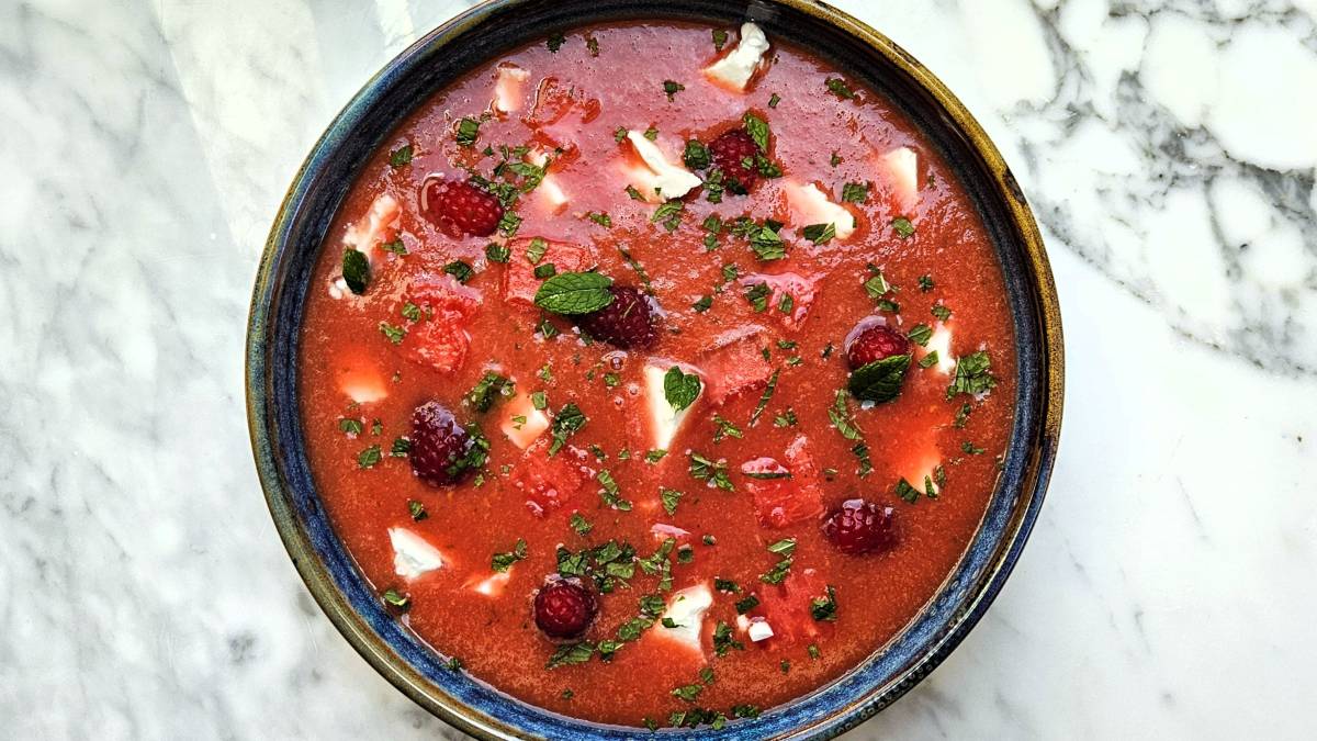 In Draufsicht: Die Wassermelonen-Himbeeren-Kaltschale. In der roten Flüssigkeit schwimmen ganze Himbeeren, einige Wassermelonenwürfel und Fetastücke. Darauf verteilt gehackte Minzblätter.