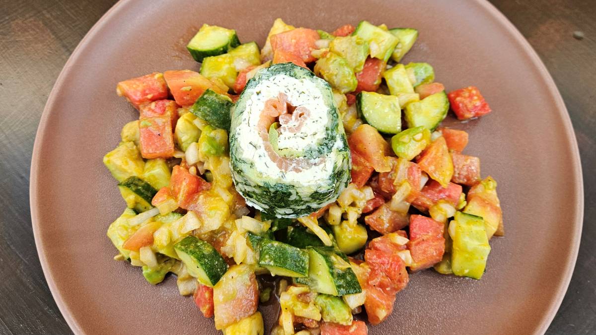 Das Bild zeigt einen Teller mit dem Wassermelonensalat mit Kurkumadressing; zu sehen sind die Zutaten wie Wassermelone, Gurke und Avocado mit dem leicht cremigen gelben Dressing. Auf dem Salat befindet sich als Topping eine Spinat-Lachs-Rolle.