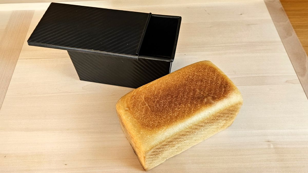 Das Bild zeigt einen Laib Weizentoastbrot aus Sauerteig auf einem Brett, daneben die leere Toastbrotform mit Deckel aus Metall.