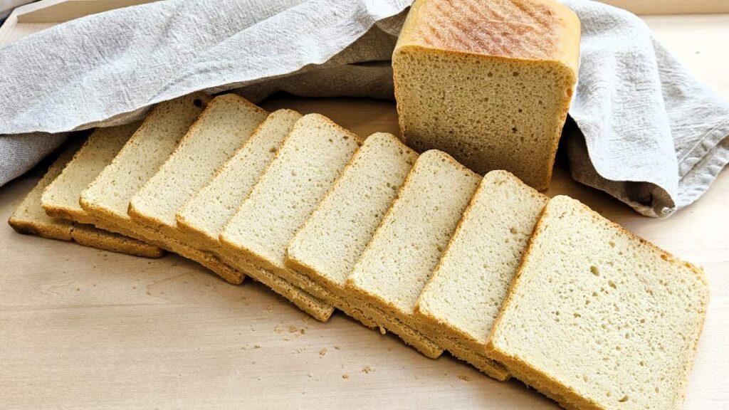 Das Bild zeigt einen Laib Toastbrot auf einem Brett, dahinter ein Tuch, davor sind Toastscheiben geschnitten angerichtet.