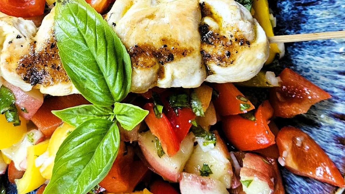 Eine Nahaufnahme vom Paprika-Pfirsich-Salat, zu sehen sind die Zutaten - Paprika, Pfirsiche, Zwiebel, Basilikum - und der Hühnerspieß mit der Marinade auf einem blauen Teller.