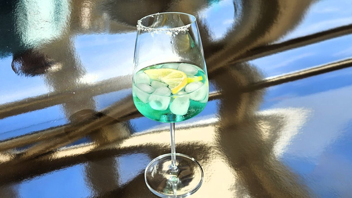 Ein einzelnes Glas mit Venturo Spritz auf einem spiegelnden Untergrund. Die türkisgrüne Farbe des Drinks ist gut erkennbar, im Drink Eiswürfel und Zitronenscheiben, auf dem halben Glasrand ist ein Salzrand.